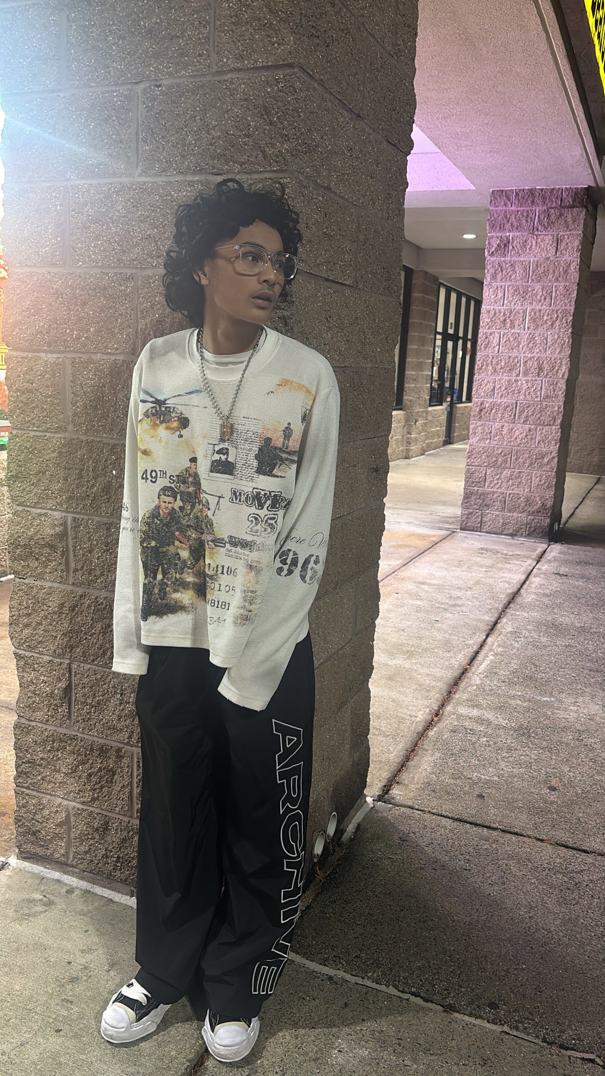 Person wearing a graphic t-shirt and dark pants leaning against a wall.